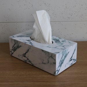 May include: White tissue box with a marble design in shades of white and green. The box is made of a hard material and has a rectangular shape.