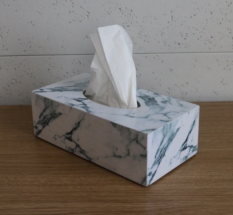 May include: White tissue box with a marble design in shades of white and green. The box is made of a hard material and has a rectangular shape.