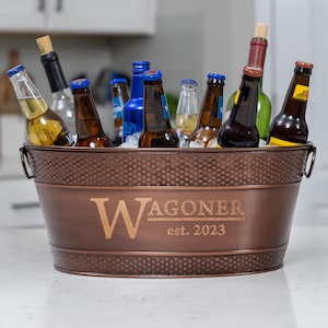 May include: A copper-colored metal beverage tub filled with ice and various bottles of beer and wine. The tub has a riveted texture and the name "WAGONER" and the year "est. 2023" are engraved on the front.