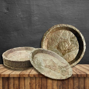 May include: A stack of brown, round, biodegradable plates made from palm leaves. The plates are arranged on a wooden surface.