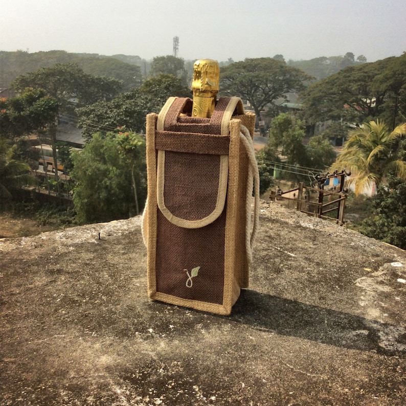 May include: A brown jute wine bottle carrier with a zippered closure and a handle. The carrier has a green leaf logo embroidered on the front.