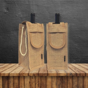 May include: Two brown burlap wine bottle carriers with a rope handle and a brown suede pocket on the front. The carriers are sitting on a wooden surface.