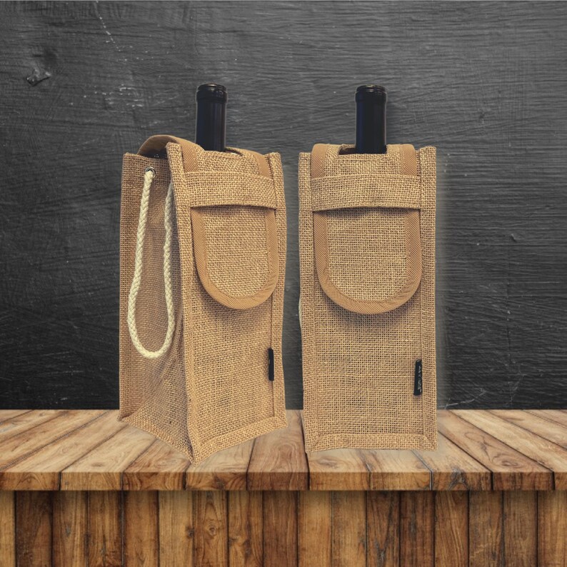 May include: Two brown burlap wine bottle carriers with a rope handle and a brown suede pocket on the front. The carriers are sitting on a wooden surface.