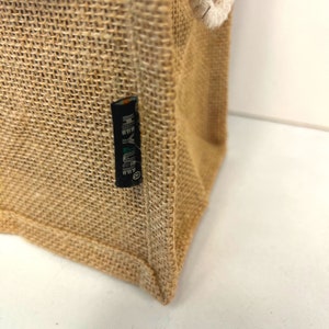 May include: Close-up of a brown burlap tote bag with a black label that reads "HEY LIFE" in white lettering.