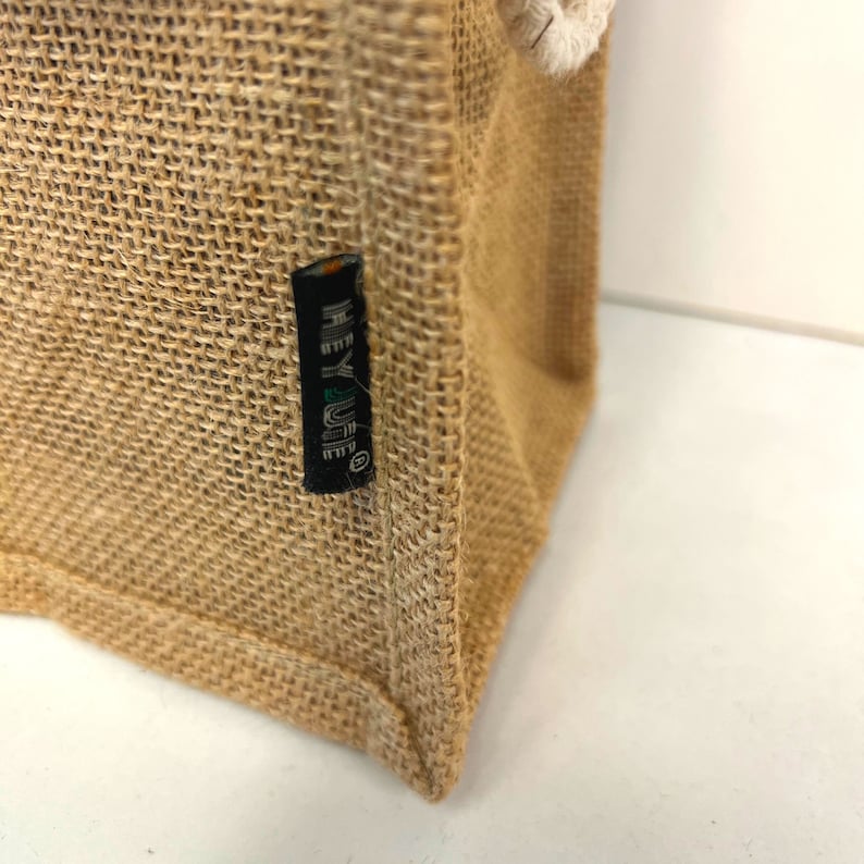 May include: Close-up of a brown burlap tote bag with a black label that reads "HEY LIFE" in white lettering.