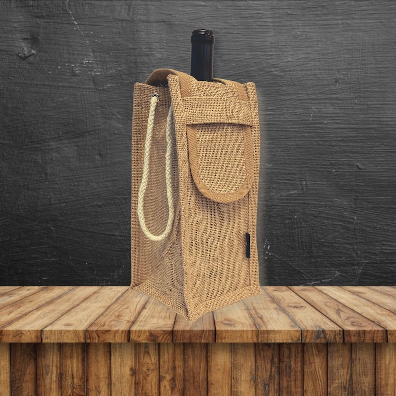 May include: A brown jute wine bottle carrier with a white rope handle and a brown fabric pocket on the front. The carrier is sitting on a wooden surface.
