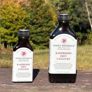 May include: Two dark brown glass bottles with black caps. The labels read "MAPLE MINERALS" and "RASPBERRY DIET CATALYST". The bottles are on a wooden surface with a blurred background of trees.