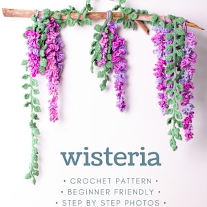 May include: A crochet wisteria wall hanging featuring three purple and pink flower clusters with green leaves suspended from a wooden branch. The text reads: wisteria, CROCHET PATTERN, BEGINNER FRIENDLY, STEP BY STEP PHOTOS.