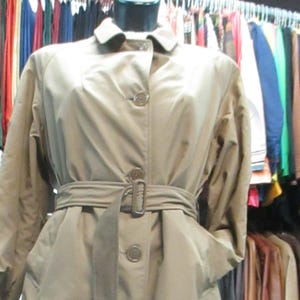 May include: A tan trench coat displayed on a mannequin. The coat features a button-up front, a belted waist with a buckle, and pockets. The coat is long-sleeved and has a collar. Clothing racks with various garments are visible in the background.