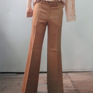 May include: A retro outfit displayed on a mannequin. It features a long-sleeved shirt with a cream and orange floral design and brown flared trousers. The shirt has a collared neckline and buttoned cuffs. The trousers have a high waist and wide legs.