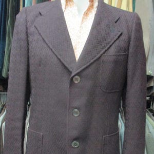 May include: A dark brown blazer with a textured pattern, three buttons, and two front pockets. The blazer is displayed on a mannequin, with a patterned shirt collar visible. A rack of other jackets is in the background.