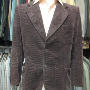 Pode incluir: Blazer de veludo cotelê marrom com padrão de espinha de peixe. O blazer tem lapelas entalhadas, dois bolsos frontais e fecho de dois botões. Uma camisa branca com gola é visível por baixo. O blazer é exibido em um manequim.