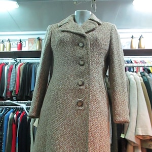 Könnte beinhalten: Ein Vintage-Mantel in Braun und Creme mit einem strukturierten Muster. Der Mantel hat einen klassischen Kragen, lange Ärmel und eine Knopfleiste vorne. Der Mantel wird auf einer Schaufensterpuppe präsentiert, die seine maßgeschneiderte Passform und sein Design zeigt.