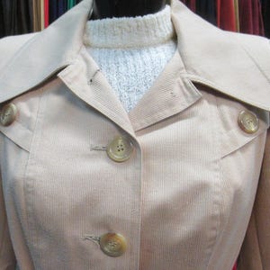 May include: A light tan corduroy jacket with a wide collar and three large, round buttons. The jacket is buttoned up, and a white sweater is visible beneath the collar. The jacket has a tailored fit and a classic design.
