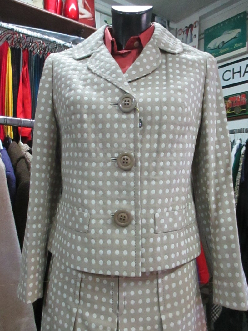 May include: A beige blazer and skirt set with white polka dots. The blazer has a notched collar and three buttons down the front. The skirt is a-line and has pleats.