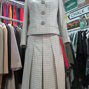 May include: A beige and white polka dot skirt suit with a button-up jacket. The skirt is pleated and the jacket has a collar and pockets.
