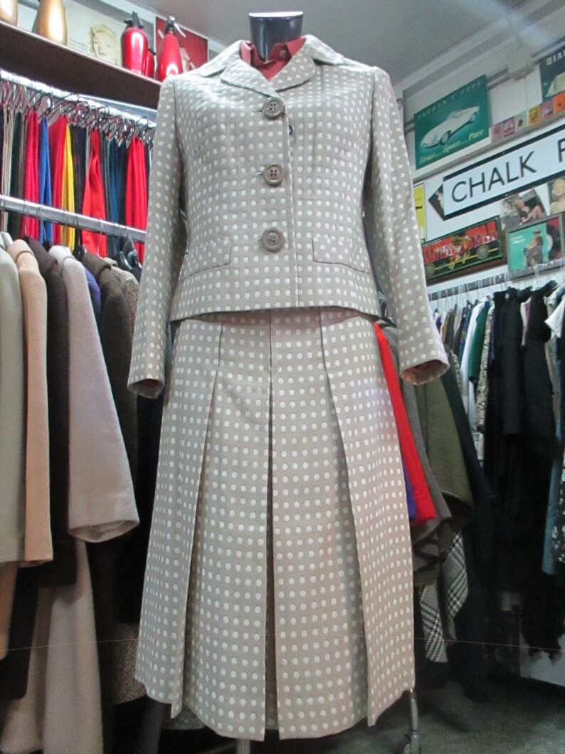 May include: A beige and white polka dot skirt suit with a button-up jacket. The skirt is pleated and the jacket has a collar and pockets.
