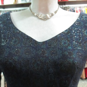 May include: A sleeveless, dark blue dress with a green and silver floral pattern. The dress has a V-neckline and is adorned with a silver rhinestone necklace.