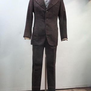 Op de afbeelding: Een bruin pak uitgestald op een mannequin. De jas heeft drie knopen, een gekerfde revers en twee voorzakken. Een wit overhemd met kraag en een stropdas met patroon zijn zichtbaar. De broek van het pak heeft rechte pijpen.