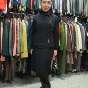 May include: A woman wearing a black turtleneck sweater and a black skirt with a matching jacket. She is standing in front of a rack of clothing and holding a black handbag.