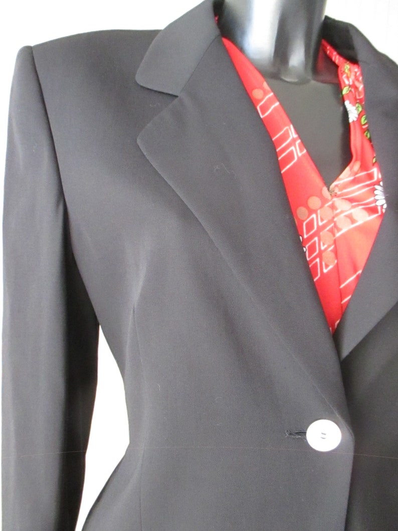 May include: A black blazer with a single white button closure. A red scarf with a white and floral pattern is visible beneath the lapel. The blazer has a classic notched collar and a tailored fit.