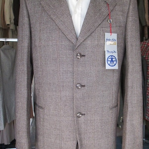 May include: A brown and white herringbone patterned blazer with a white collared shirt underneath. The blazer has a three-button closure and a pocket on the left side. The blazer has a tag that reads "Jake Solo" and "Trascendent".