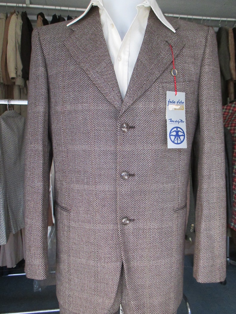 May include: A brown and white herringbone patterned blazer with a white collared shirt underneath. The blazer has a three-button closure and a pocket on the left side. The blazer has a tag that reads "Jake Solo" and "Trascendent".