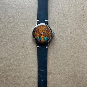 Puede incluir: Un reloj de pulsera con caja plateada y correa de cuero azul oscuro. La esfera del reloj presenta un dise&ntilde;o detallado de &aacute;rbol en tonos naranja, dorado y turquesa. La correa mide aproximadamente 23 cm de largo.