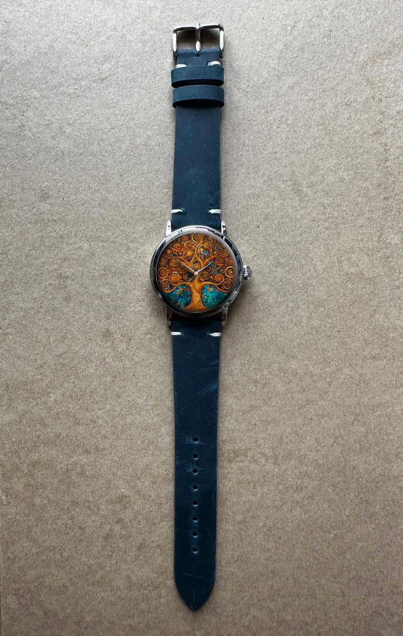 Puede incluir: Un reloj de pulsera con caja plateada y correa de cuero azul oscuro. La esfera del reloj presenta un dise&ntilde;o detallado de &aacute;rbol en tonos naranja, dorado y turquesa. La correa mide aproximadamente 23 cm de largo.