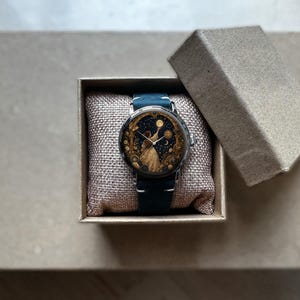 May include: A round wristwatch with a dark blue leather band and a celestial-themed face featuring a figure and stars. The watch is presented in a small, open, beige box with a textured interior, suggesting a gift or display item.