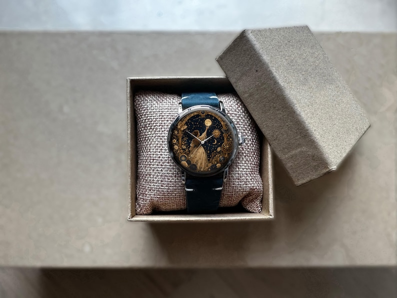 May include: A round wristwatch with a dark blue leather band and a celestial-themed face featuring a figure and stars. The watch is presented in a small, open, beige box with a textured interior, suggesting a gift or display item.