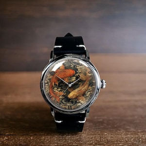 Puede incluir: Reloj de pulsera redondo con correa de cuero negro y caja plateada. La esfera del reloj presenta dos peces koi en tonos naranja y dorado, sobre un fondo detallado. El reloj tiene un diseño clásico.