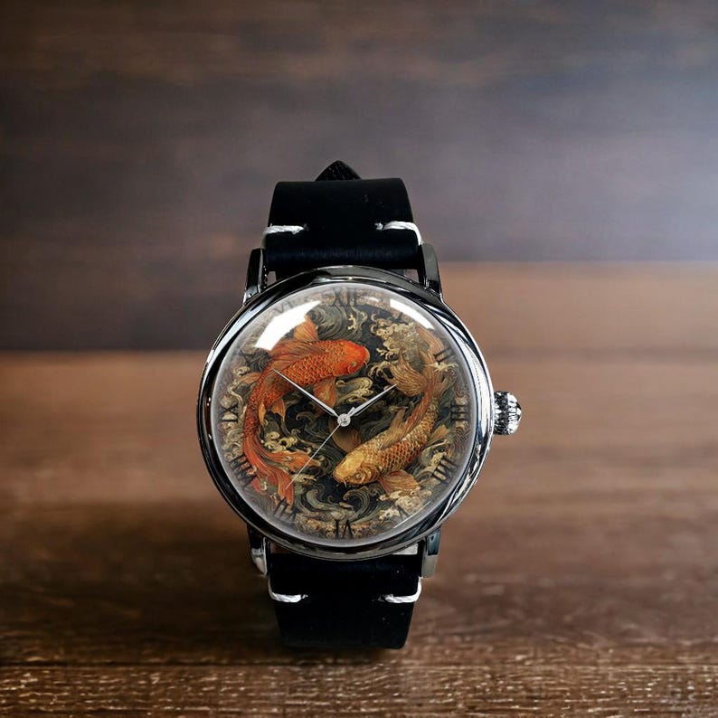 May include: A round wristwatch with a black leather band and a silver-colored case. The watch face features two koi fish in shades of orange and gold, set against a detailed background. The watch has a classic design.