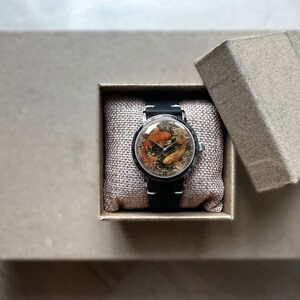 May include: A wristwatch with a black band and a round face featuring a koi fish design in shades of orange, brown, and gold. The watch is presented in an open, square, cardboard gift box with a linen-like cushion.