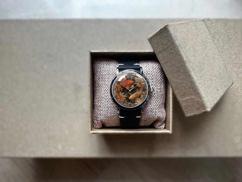 May include: A wristwatch with a black band and a round face featuring a koi fish design in shades of orange, brown, and gold. The watch is presented in an open, square, cardboard gift box with a linen-like cushion.