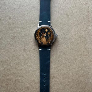 May include: A silver-toned wristwatch with a dark blue leather strap. The watch face features a celestial design with a figure reaching for the moon and stars. The strap is approximately 23 cm long.