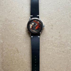 Op de afbeelding: Een polshorloge met een zwarte leren band en een ronde wijzerplaat. De wijzerplaat heeft een koi-karperontwerp in rood, wit en zwart. Het horloge heeft een zilverkleurige kast en een gespsluiting.