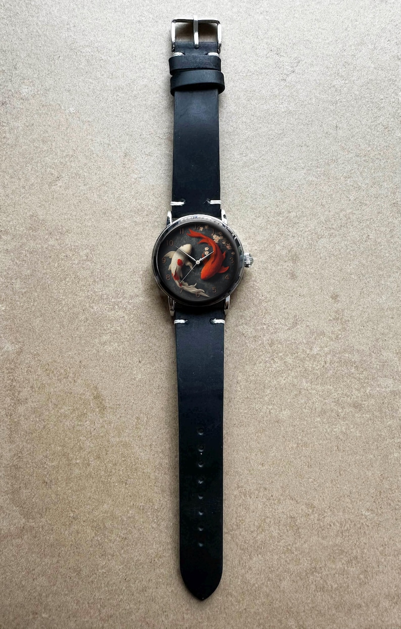 Op de afbeelding: Een polshorloge met een zwarte leren band en een ronde wijzerplaat. De wijzerplaat heeft een koi-karperontwerp in rood, wit en zwart. Het horloge heeft een zilverkleurige kast en een gespsluiting.