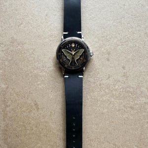 May include: A black leather watch with a silver case and a detailed face featuring a moth design. The watch has a black band with white stitching and a silver buckle. The watch face also has moon phases and Roman numerals.
