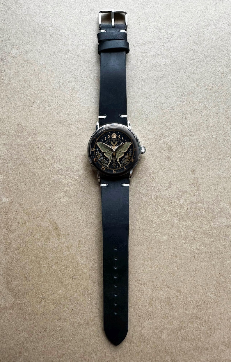 May include: A black leather watch with a silver case and a detailed face featuring a moth design. The watch has a black band with white stitching and a silver buckle. The watch face also has moon phases and Roman numerals.