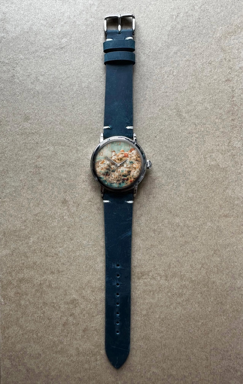 Pode incluir: Um rel&oacute;gio de pulso com uma pulseira de couro azul e uma caixa prateada. O mostrador do rel&oacute;gio apresenta uma ilustra&ccedil;&atilde;o colorida de uma paisagem urbana. A pulseira tem cerca de 24 cm de comprimento.