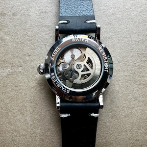Puede incluir: Primer plano de un reloj de acero inoxidable con correa de cuero negro. La esfera del reloj es transparente, revelando el intrincado movimiento mec&aacute;nico. Las palabras "Water Resistant" y "Stainless Steel" son visibles.