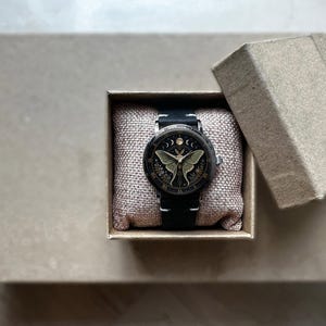 May include: A black wristwatch with a detailed face featuring a moth design, presented in a small, open cardboard box. The watch has a black leather band and is set on a textured, beige fabric.