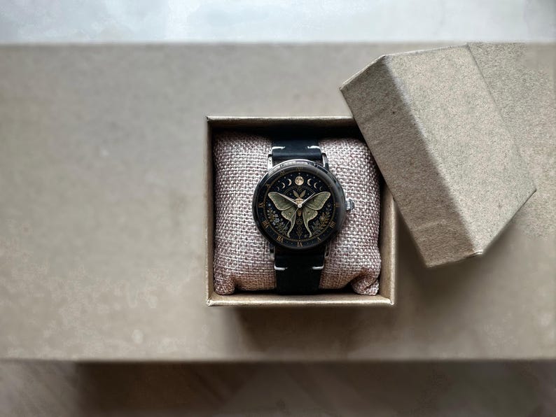 May include: A black wristwatch with a detailed face featuring a moth design, presented in a small, open cardboard box. The watch has a black leather band and is set on a textured, beige fabric.