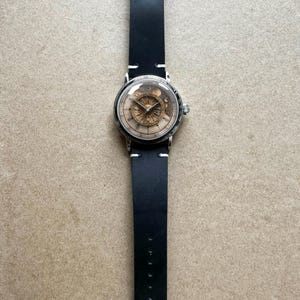 Puede incluir: Un reloj de pulsera plateado con una correa de cuero negro. La esfera del reloj tiene una cubierta transparente, que revela un mecanismo detallado de color dorado. La correa negra est&aacute; cosida con hilo blanco.