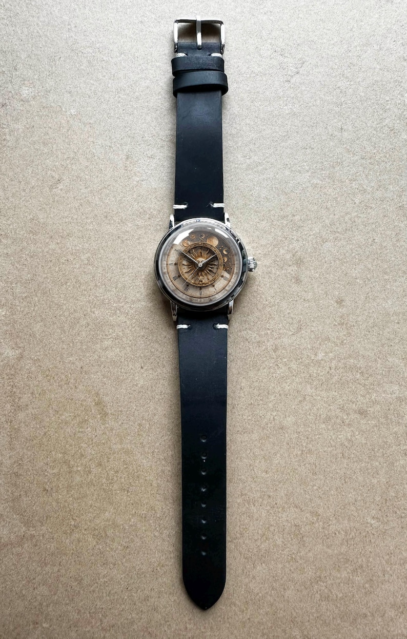 Puede incluir: Un reloj de pulsera plateado con una correa de cuero negro. La esfera del reloj tiene una cubierta transparente, que revela un mecanismo detallado de color dorado. La correa negra est&aacute; cosida con hilo blanco.