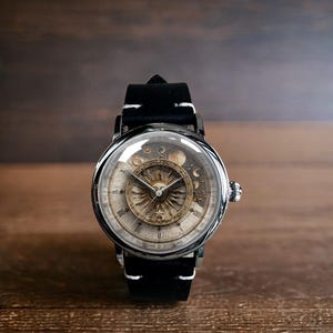 Puede incluir: Un reloj de pulsera redondo con una correa de cuero negro y costuras blancas. La esfera del reloj presenta un dise&ntilde;o de sol y luna con n&uacute;meros romanos. El reloj tiene una caja plateada y una cubierta de cristal transparente.