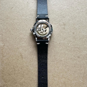 Puede incluir: Un reloj de cuero negro con caja de acero inoxidable y esfera esqueleto. El reloj tiene una correa de cuero negro y una hebilla plateada. La esfera del reloj es visible, mostrando el mecanismo interno. El reloj es resistente al agua.