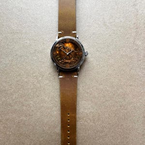 May include: A wristwatch with a brown leather band and a silver-colored case. The watch face features a detailed illustration of a dinosaur skeleton in a sepia tone. The band has a classic buckle closure and is approximately 1 inch wide.