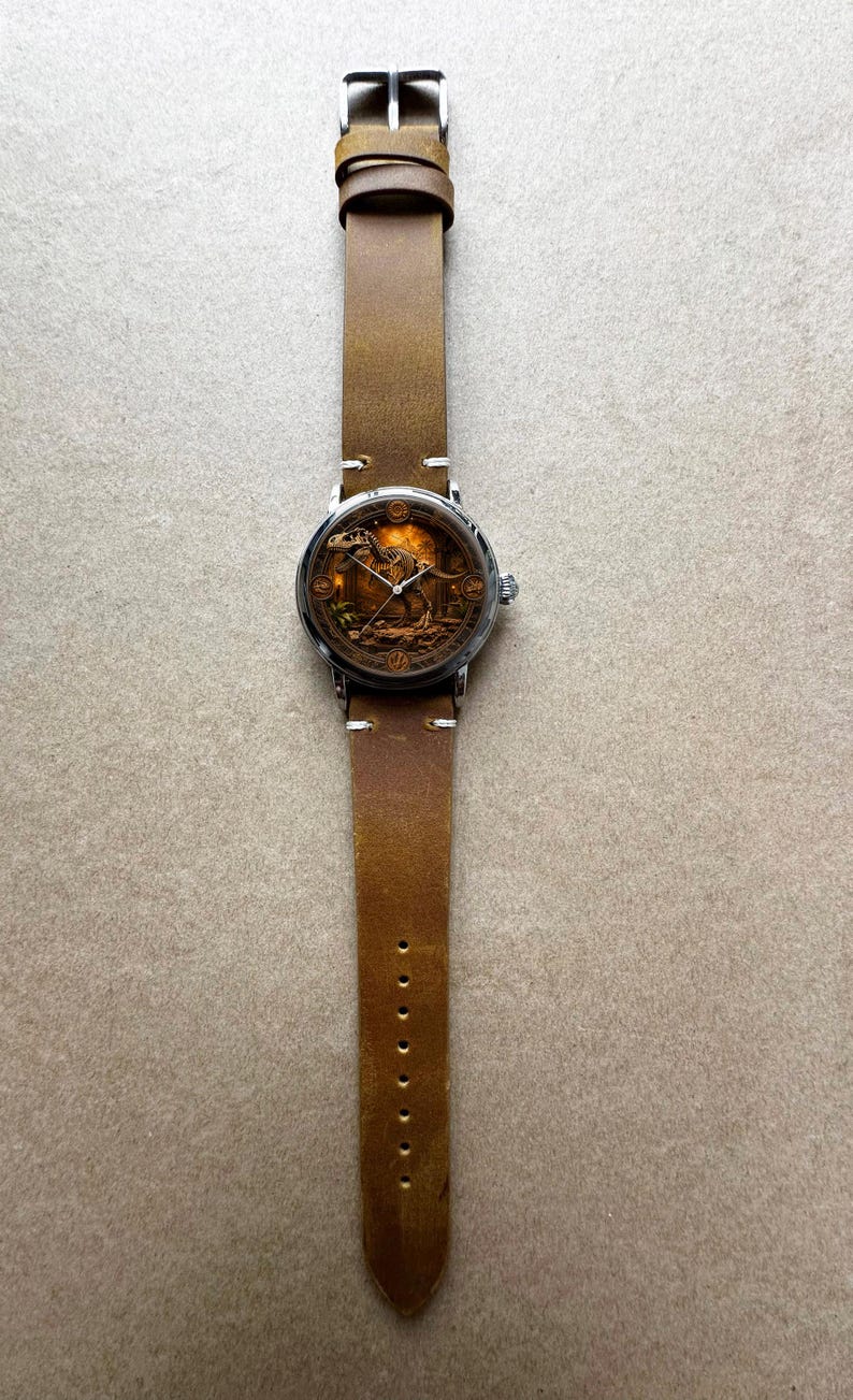 May include: A wristwatch with a brown leather band and a silver-colored case. The watch face features a detailed illustration of a dinosaur skeleton in a sepia tone. The band has a classic buckle closure and is approximately 1 inch wide.